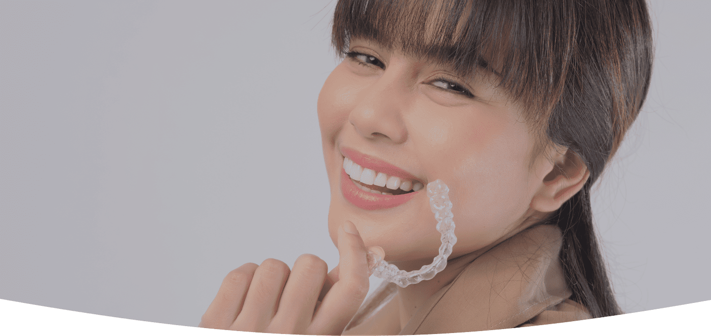 A smiling young woman, who appears to be of East Asian descent based on her features, holds a clear aligner. Limestone Hills Orthodontics in Austin, TX, offers clear aligner treatments.