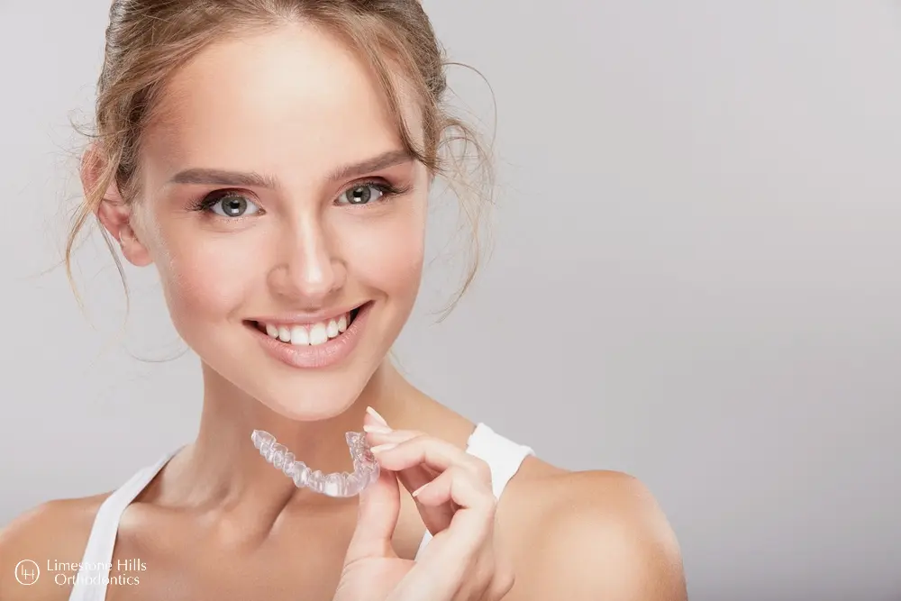 A smiling young woman, who appears to be of Caucasian descent based on her features and complexion, holds clear aligners, possibly Invisalign. Limestone Hills Orthodontics in Austin, TX.