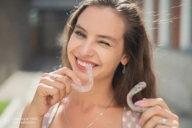 a woman smiling to the camera holding two Invisaligns. Does Invisalign Hurt?