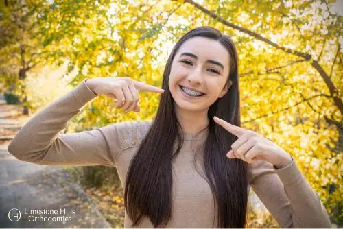 A smiling young woman, who appears to be of Caucasian descent based on her features and complexion, points to her braces outdoors. Limestone Hills Orthodontics in Austin, TX.