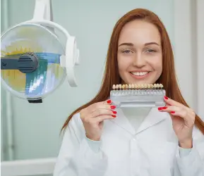 A woman in a white lab coat holding up a dental shade guide and smiling at the camera. The image is taken in a dental clinic setting with dental equipment visible in the background. The clinic name is Limestone Hills Orthodontics and the location is Austin, TX.