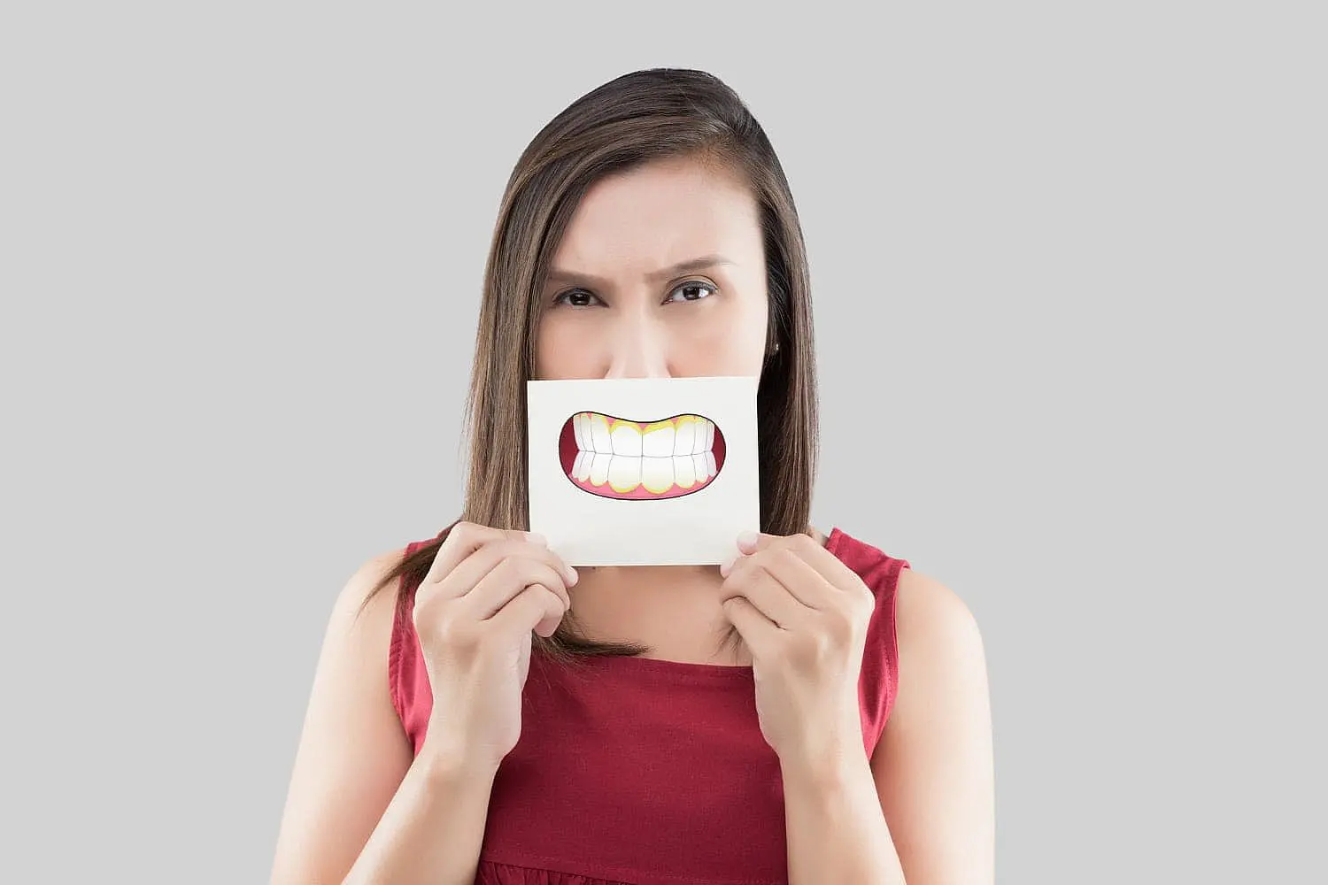 A woman with a neutral expression holds a drawing of teeth with a noticeable underbite in front of her mouth. This image could subtly suggest the genetic factors that can contribute to the development of an underbite, a condition that Limestone Hills Orthodontics in Austin, TX, can address.