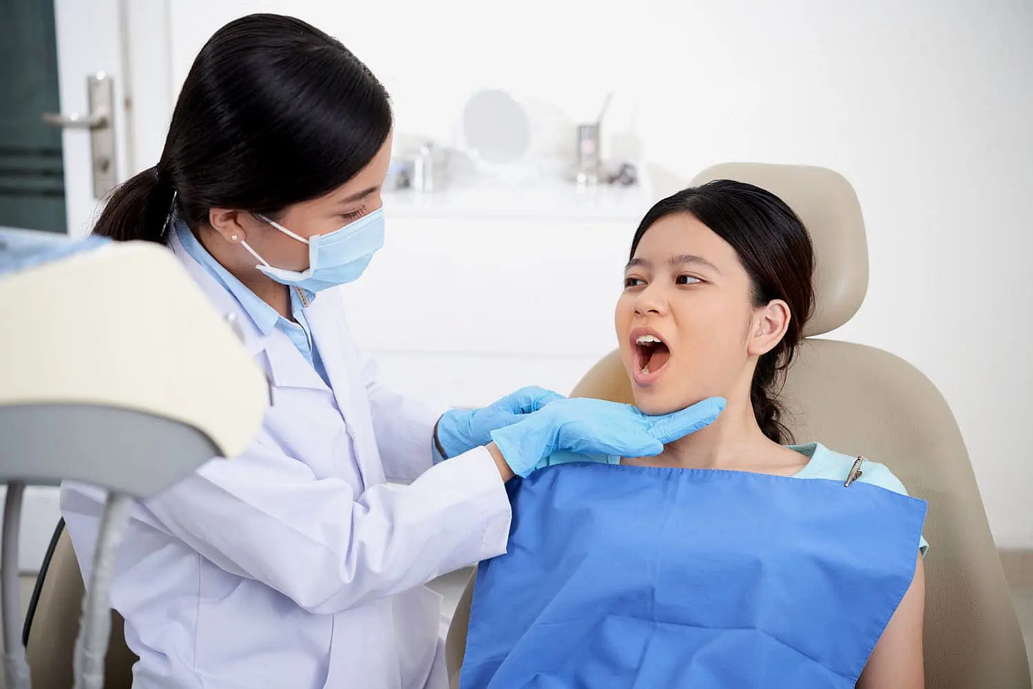 A dentist, who appears to be of Asian descent based on her features, wearing a face mask and blue gloves, is examining the open mouth of a young patient, who also appears to be of Asian descent, seated in a dental chair. This scene could represent an initial orthodontic consultation at Limestone Hills Orthodontics in Austin, TX, to assess the patient's oral health.