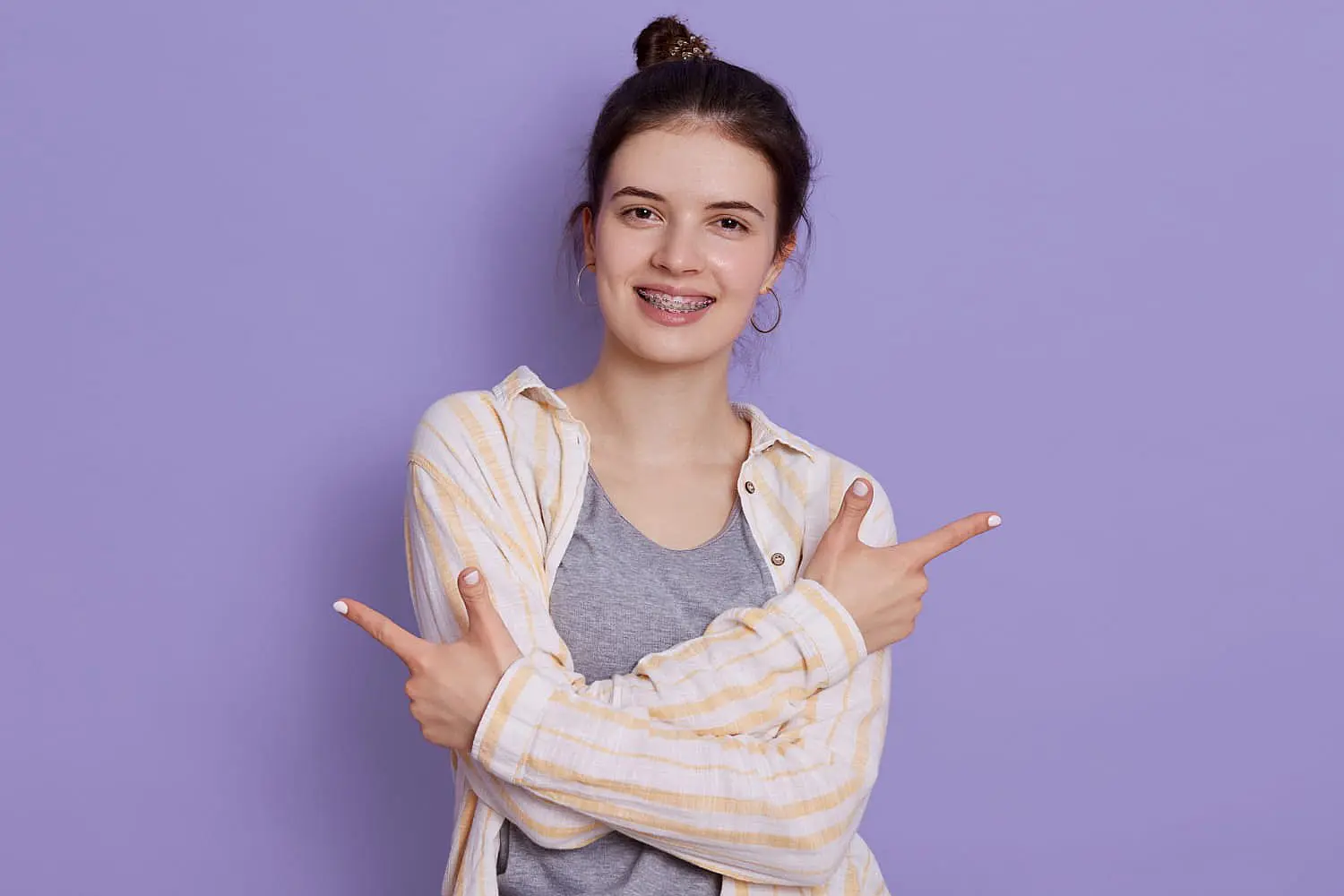 A smiling young woman with braces points with both hands in opposite directions against a purple background. This image could represent the various factors that can influence dental alignment, including potential environmental factors contributing to conditions like an underbite, which can be assessed and treated at Limestone Hills Orthodontics in Austin, TX.