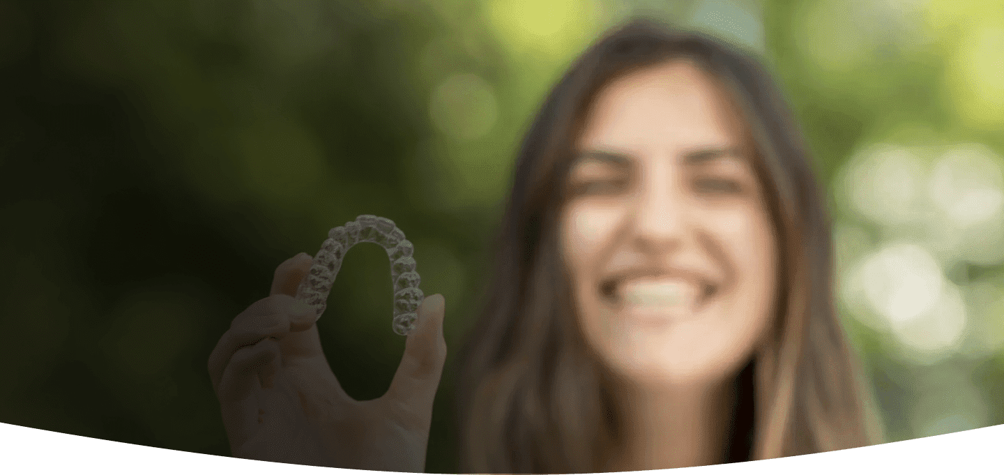 A blurred smiling woman, who appears to be of Caucasian descent based on her features and complexion, holds clear aligners. Limestone Hills Orthodontics in Austin, TX, offers Invisalign.