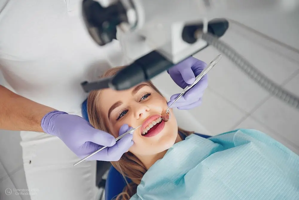 A dental professional, whose race and ethnicity are not discernible from the image, examines the braces of a smiling young woman, who appears to be of Caucasian descent based on her features and complexion. Limestone Hills Orthodontics in Austin, TX.