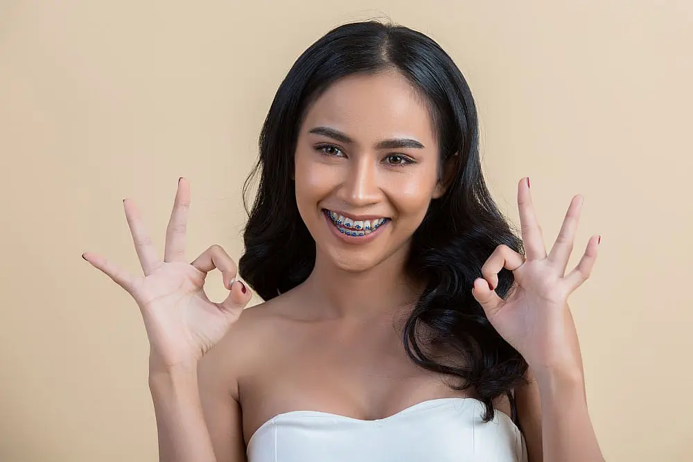 A smiling young woman, who appears to be of Southeast Asian descent based on her features and complexion, shows her braces and makes "okay" signs. Limestone Hills Orthodontics in Austin, TX.
