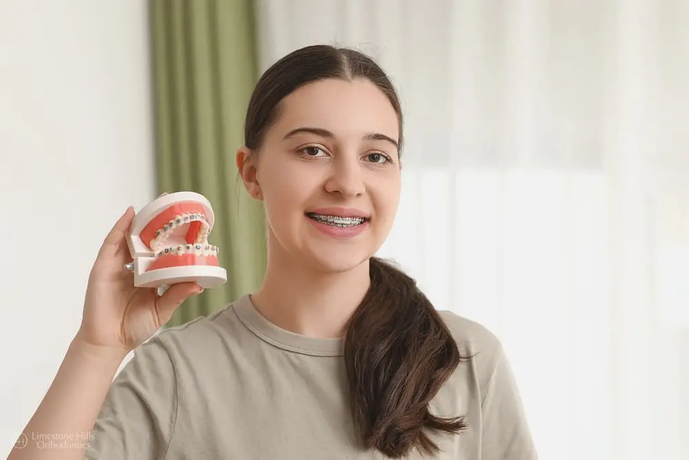 A young woman with braces holds a model of teeth with braces in Austin, TX. She is smiling slightly.