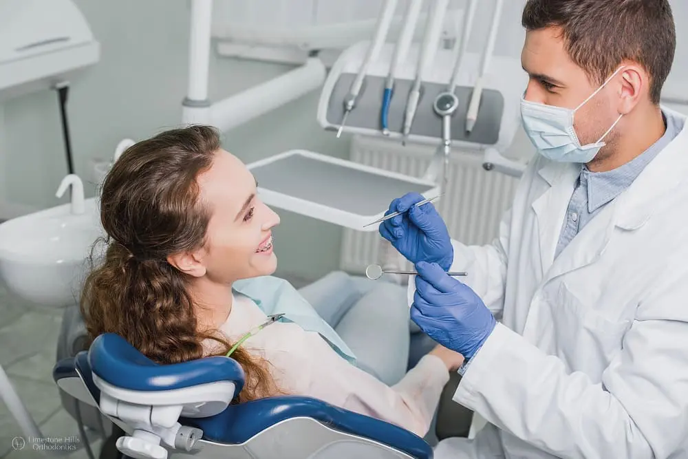Orthodontist wearing mask and latex gloves standing next to a smiling woman with metal braces during a dental check-up for her broken braces bracket.