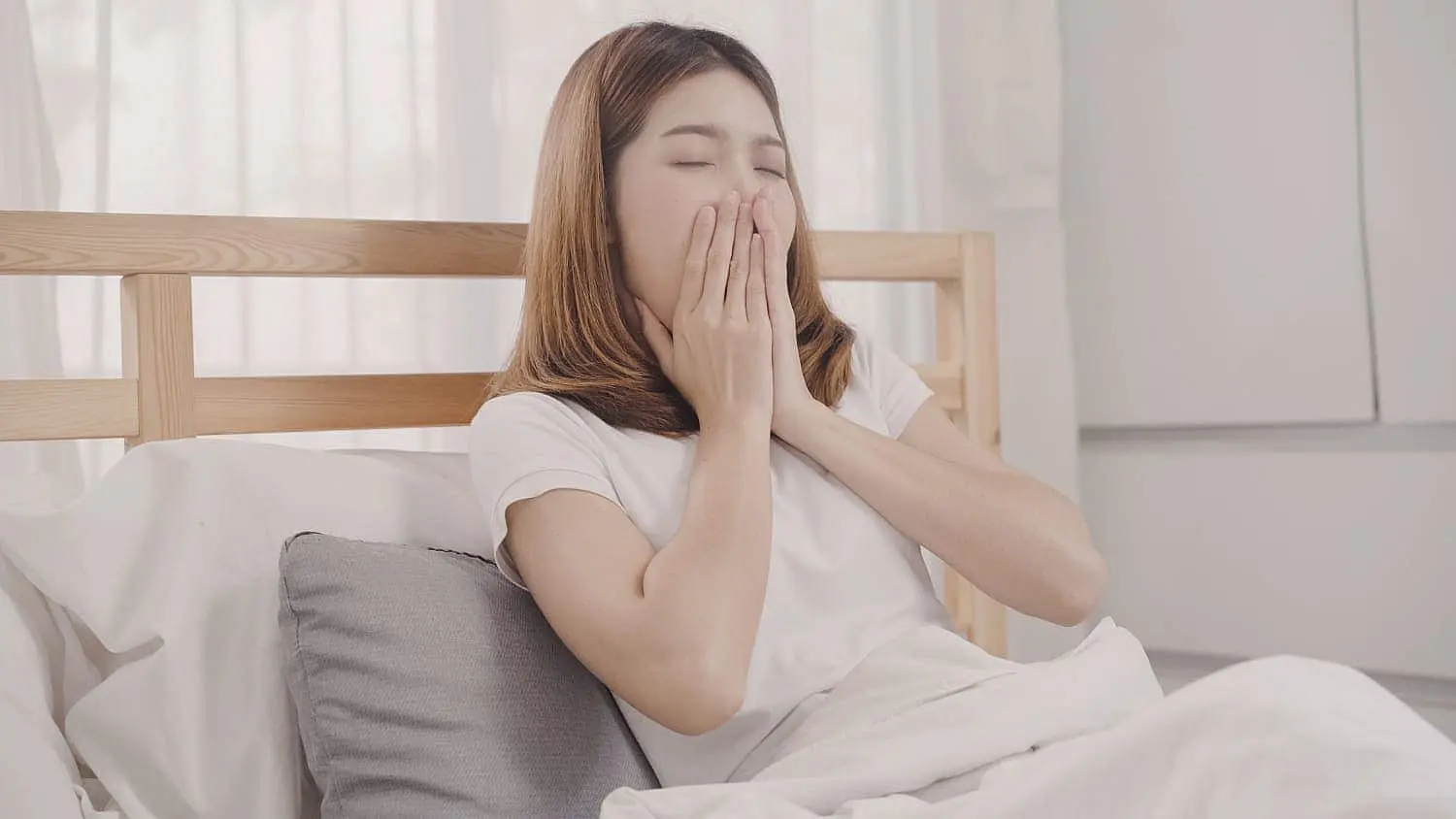 A young woman, who appears to be of Asian descent based on her features, is sitting in bed and yawning, with her eyes closed and hands covering her mouth. This image could subtly relate to the topic of sleep and breathing, which can sometimes have implications for oral health. Limestone Hills Orthodontics in Austin, TX, can provide guidance on the relationship between oral habits and orthodontic health.