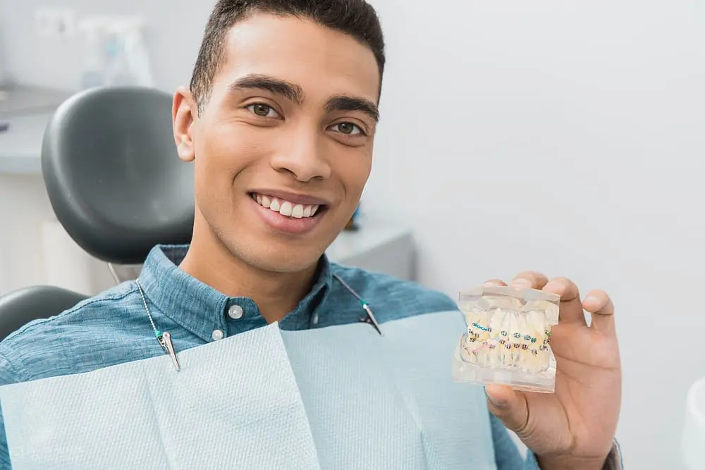 Smiling patient in blue bib holding a dental model with metal braces illustrating "can braces stain my teeth?"