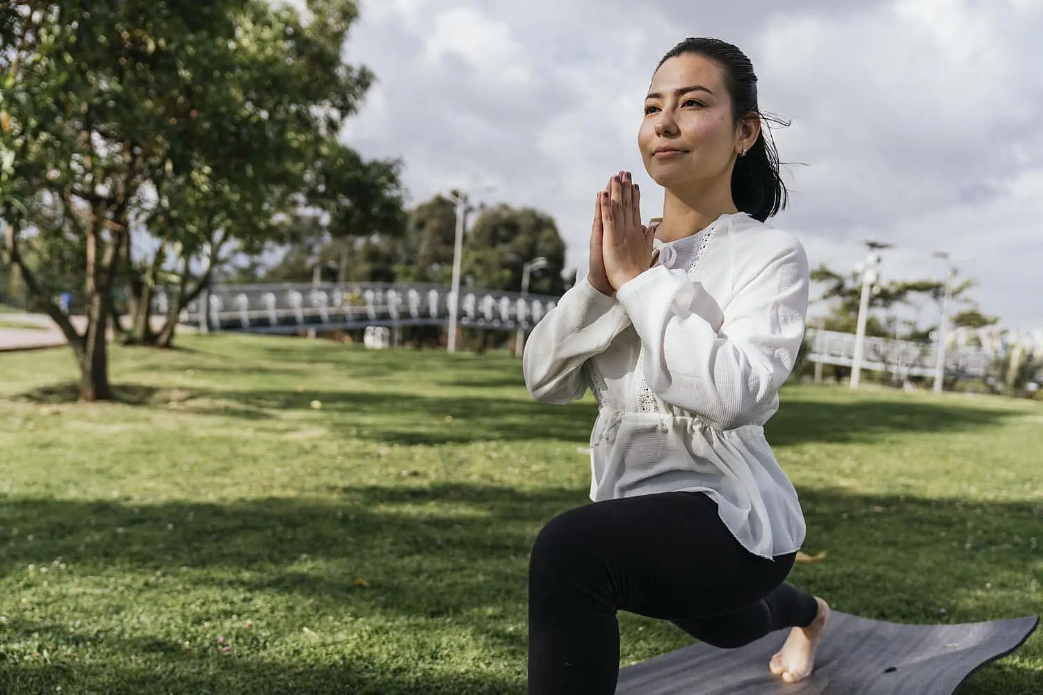 A young woman, who appears to be of Asian descent based on her facial features and hair, is practicing yoga outdoors on a mat in a park-like setting. Her hands are pressed together in a prayer position in front of her chest, and she is in a lunge pose with her gaze directed upwards. While seemingly unrelated to orthodontics, Limestone Hills Orthodontics in Austin, TX, recognizes the importance of overall well-being for their patients.
