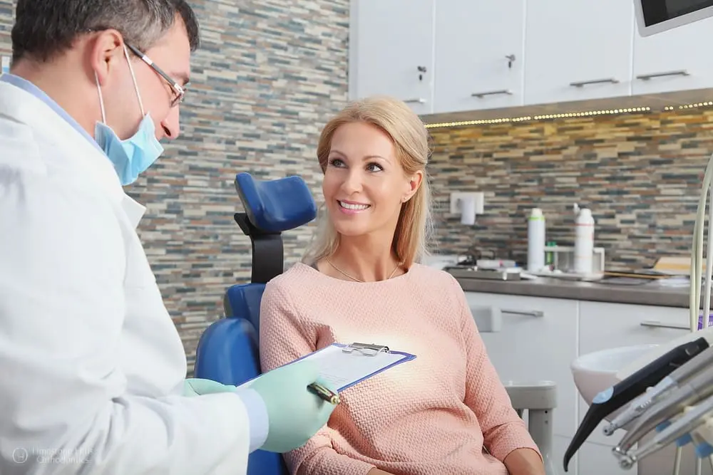 Orthodontist wearing gloves and holding a clipboard while discussing treatment with a smiling female patient as one of the orthodontic consultation process.