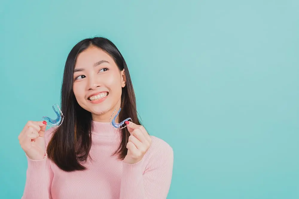 Portrait of a young Asian woman smiling while holding silicone orthodontic retainers against a blue background after learning about the Importance of Wearing a Retainer