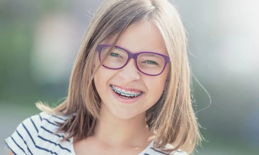 A smiling young girl, who appears to be of Caucasian descent based on her features and complexion, wears glasses and braces. Limestone Hills Orthodontics in Austin, TX, provides children's orthodontics.