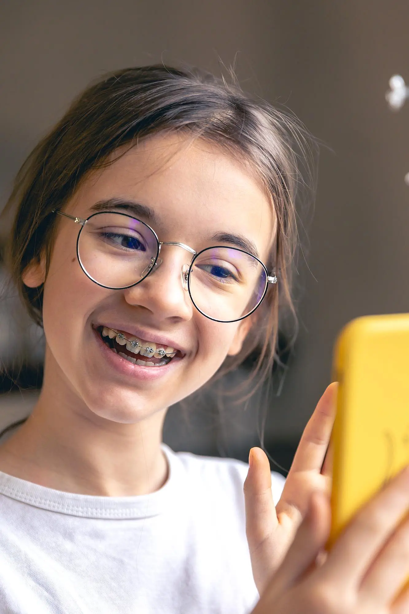 A smiling young person, who appears to be female and may be of Caucasian descent based on their features, wears glasses and braces while looking at a phone. Limestone Hills Orthodontics in Austin, TX.