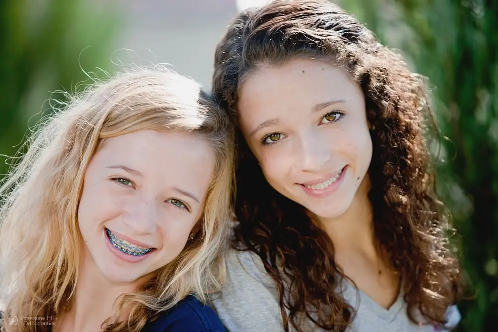 Two smiling Caucasian teenage girls with metal braces posing together in the park after applying holiday tips for braces