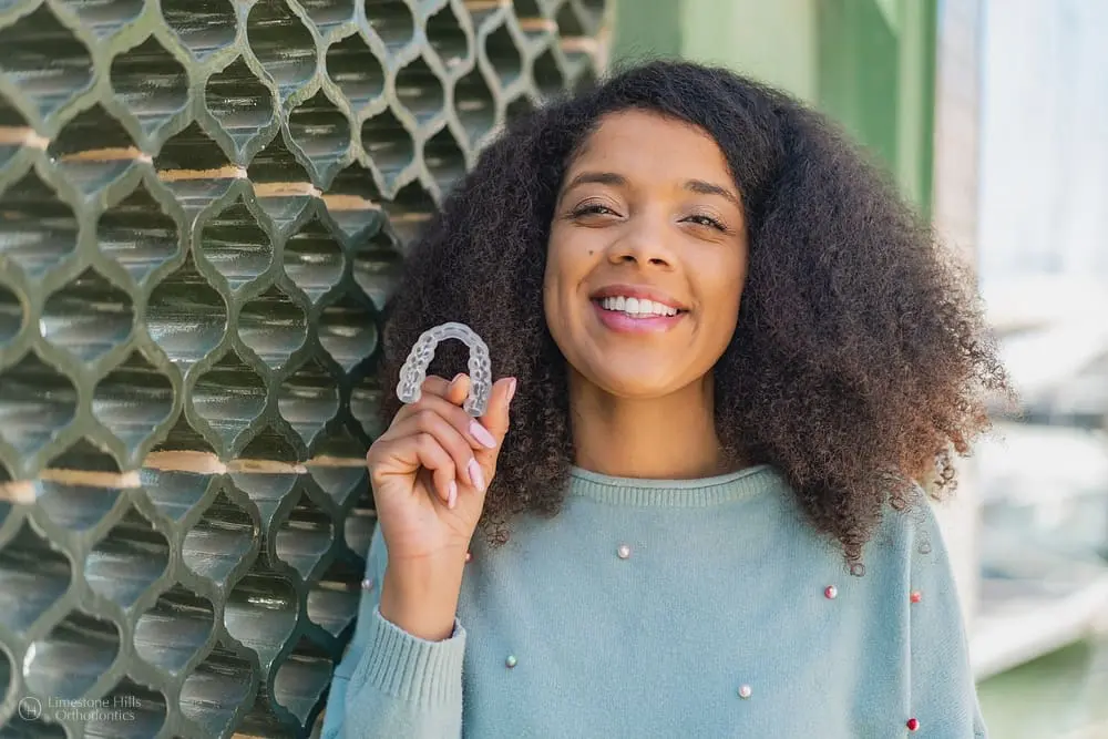 Young African American woman outdoors, holding Invisalign clear aligners and smiling trying to debunk invisalign myths