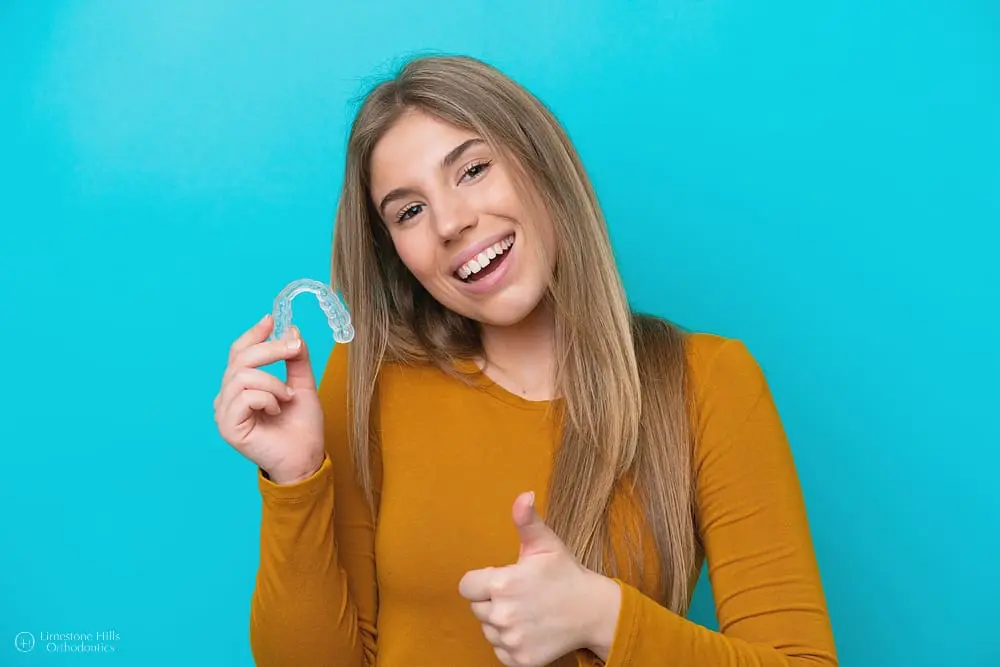 Happy woman in an orange sweater holding clear aligners against a blue background after not worrying about getting invisalign with missing teeth