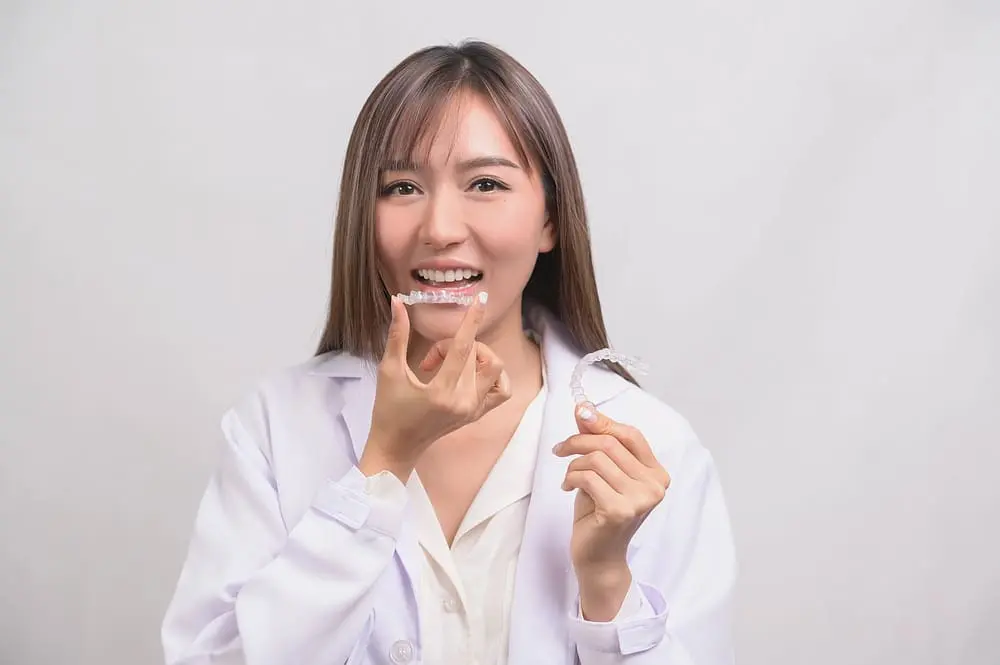 A smiling young woman, who appears to be of Asian descent based on her features, holds and points to clear aligners. Limestone Hills Orthodontics in Austin, TX, offers these.