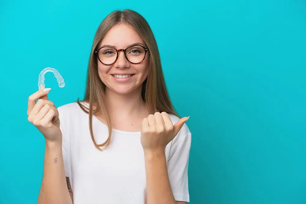 Woman wearing glasses holding Invisalign clear aligners against a blue background. after wondering about "What Happens If You Skip Wearing Invisalign?"