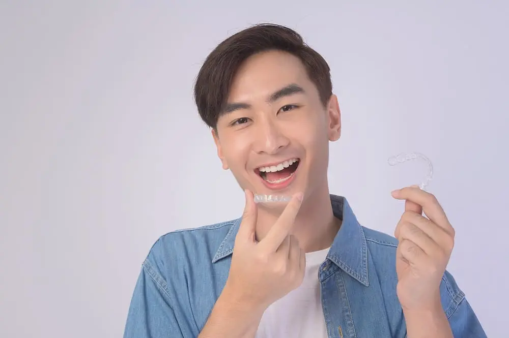 Young man in a light blue shirt smiling while holding clear aligners after not worrying about getting invisalign with missing teeth