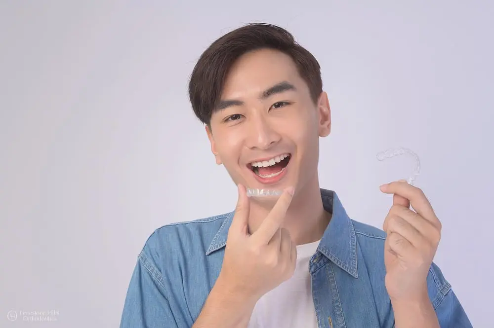 Young man in a light blue shirt smiling while holding clear aligners after not worrying about getting invisalign with missing teeth