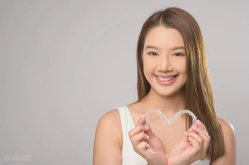 Young woman forming a heart shape with her hands while holding Invisalign clear aligners after wondering about "What Happens If You Skip Wearing Invisalign?"