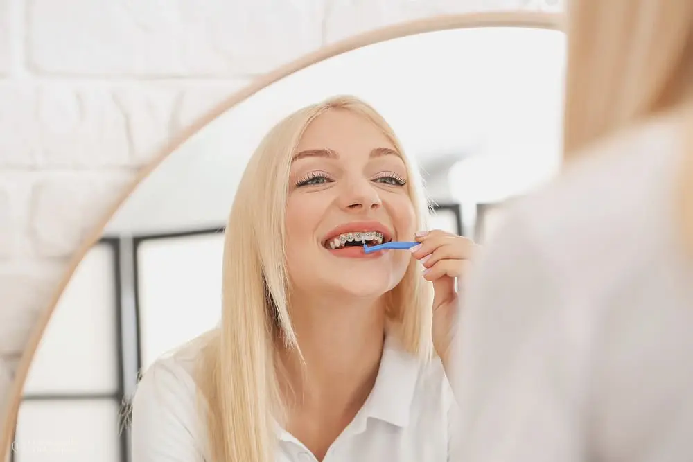 Young woman with metal braces holding a toothbrush at home, smiling confidently not worrying about her broken braces bracket