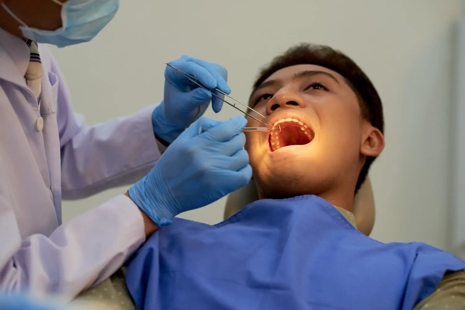 A dentist, who appears to be of Asian descent based on visual cues, wearing a surgical mask and blue gloves, examines the open mouth of a patient, who also appears to be of Asian descent, using dental instruments under a bright light. This scene could represent a consultation or examination at Limestone Hills Orthodontics in Austin, TX, to determine suitable treatment options for an underbite.