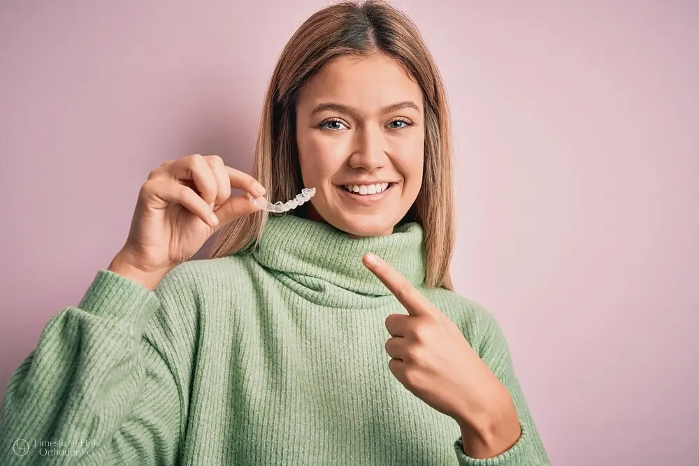 Teen girl in green sweater holding and pointing to clear aligners against a pink background, promoting Invisalign use after understanding does insurance cover invisalign in Austin, TX.
