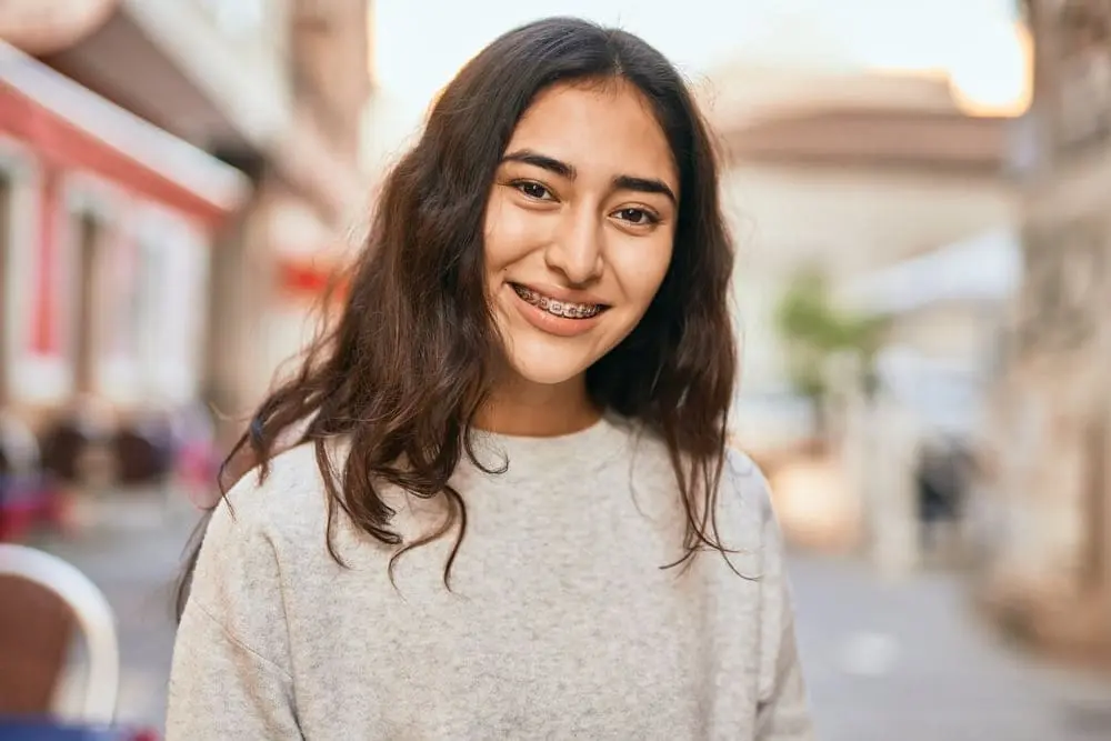 A young woman with long dark hair smiling outdoors while wearing metal braces, representing choosing the right orthodontic treatment. Limestone Hills Orthodontics in Austin, TX, helps patients find the best braces option.