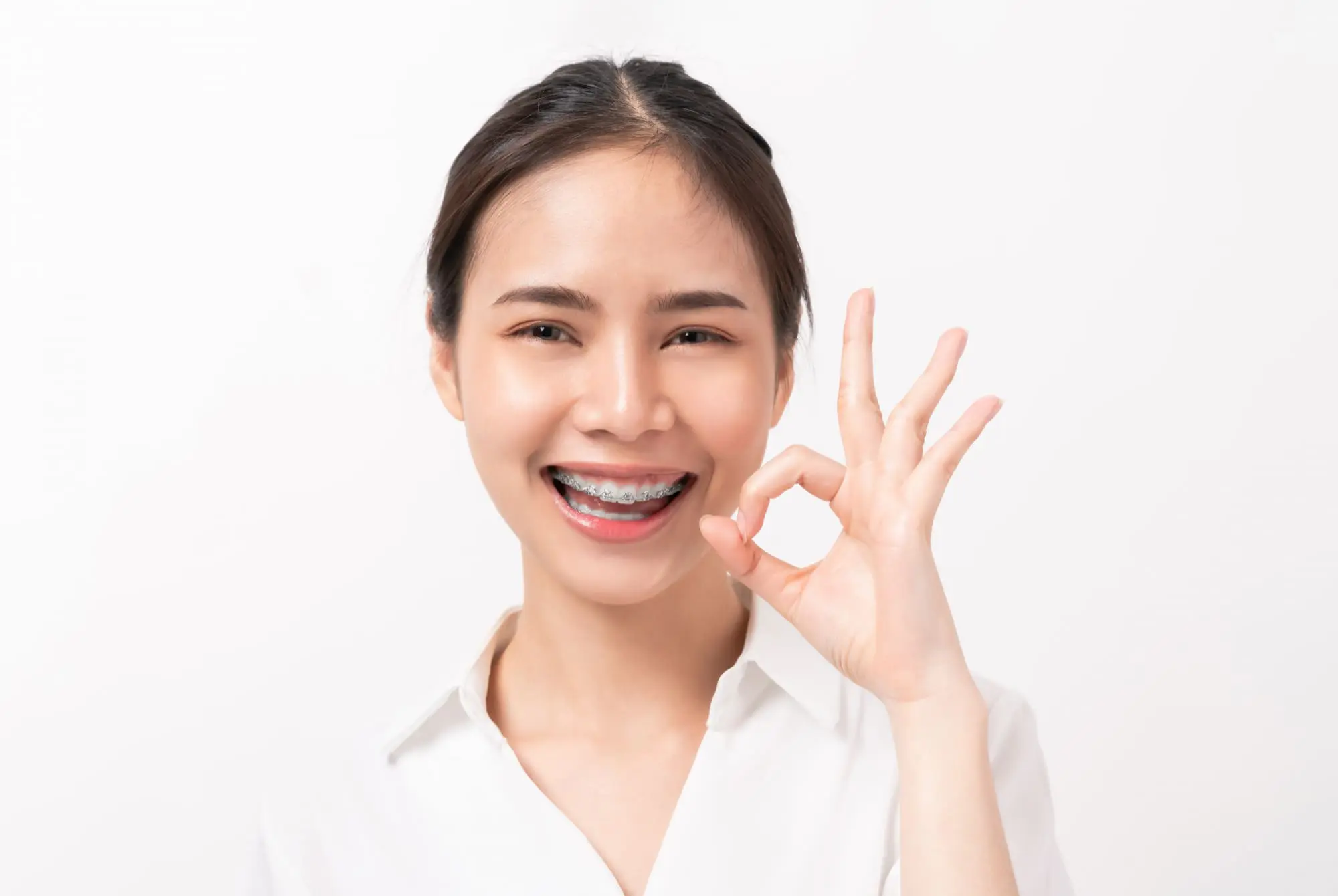 A young woman with ceramic braces smiles and gives an "OK" sign, representing Limestone Hills Orthodontics in Austin, TX.