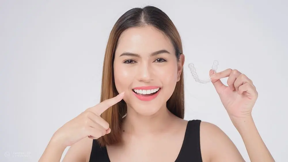 Smiling young woman holding clear aligners and pointing to teeth. Limestone Hills Orthodontics in Austin, TX, in-network care.