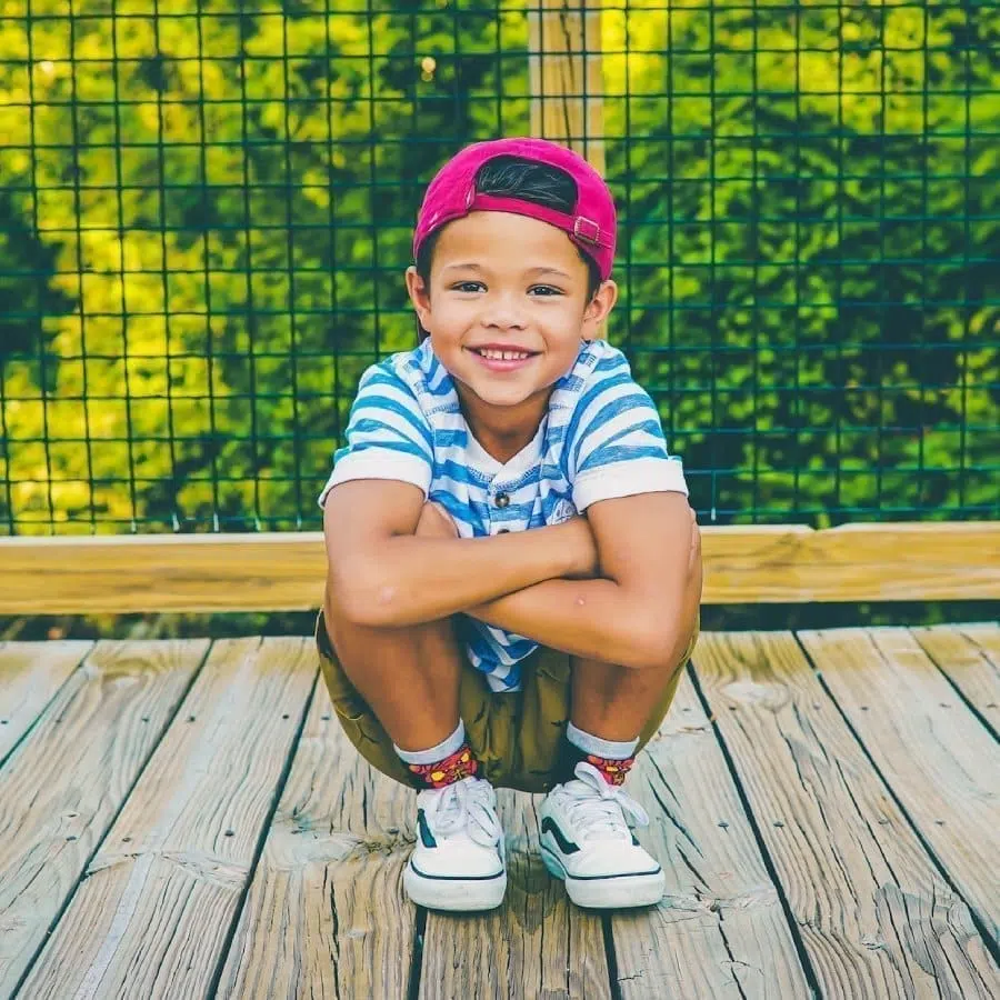 A child wearing a striped shirt, red cap, and sneakers crouches on a wooden deck. A wire fence and greenery are in the background.